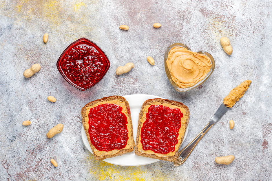 Tostadas con mermelada de frutilla sobre un plato y un pocillo con mantequilla de maní sobre una mesa gris.